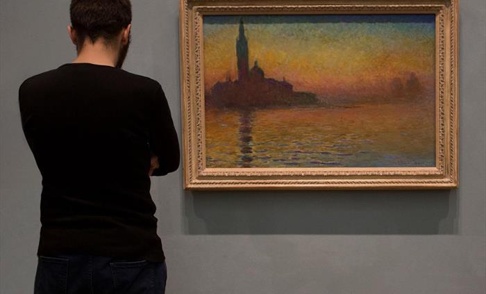 Young man with his back to the camera. He's looking at a painting showing the silhouette of a church. the sky and the reflection of the church in the